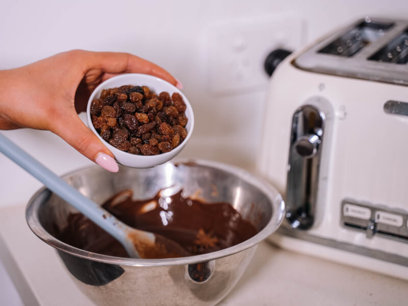 A hand pouring raisins into a bowl of melted chocolate.